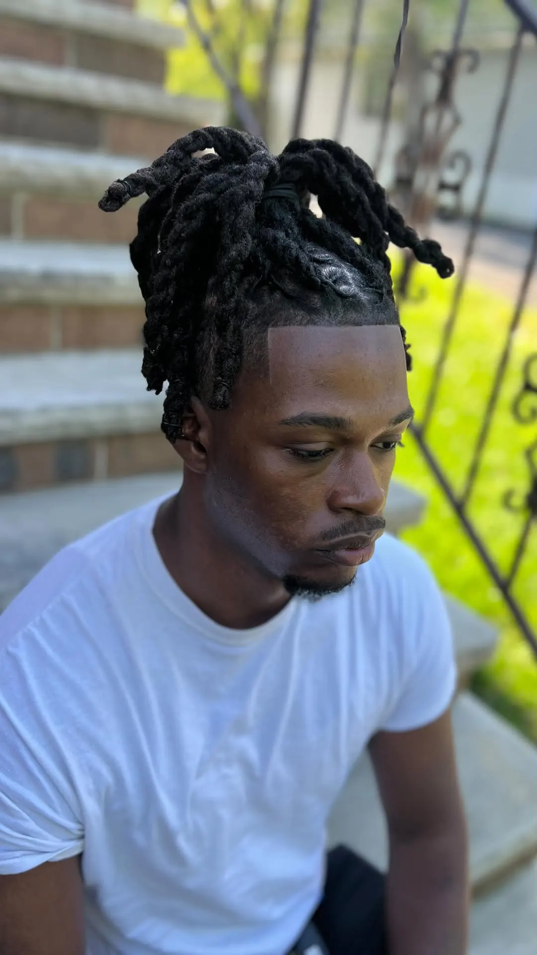 Locs gathered into a top bun over a shaved taper, outdoor portrait.